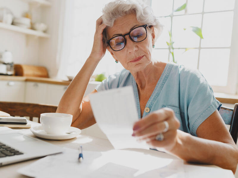 puzzled senior woman with gray hair in glasses looking at a bill holding head in hand, upset with high pricing